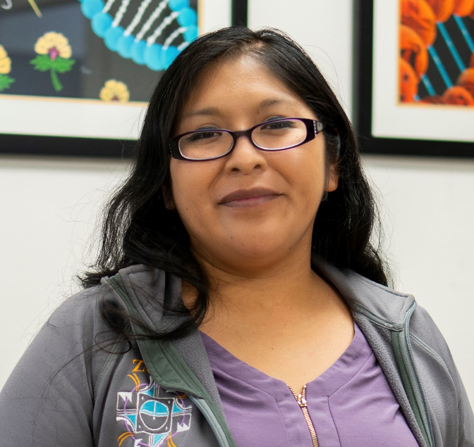 Headshot of Mallery Quetawki standing in front of two of her DNA paintings (partial views)